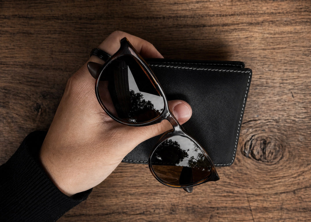 Hand holding sunglasses next to a black wallet on a wooden surface